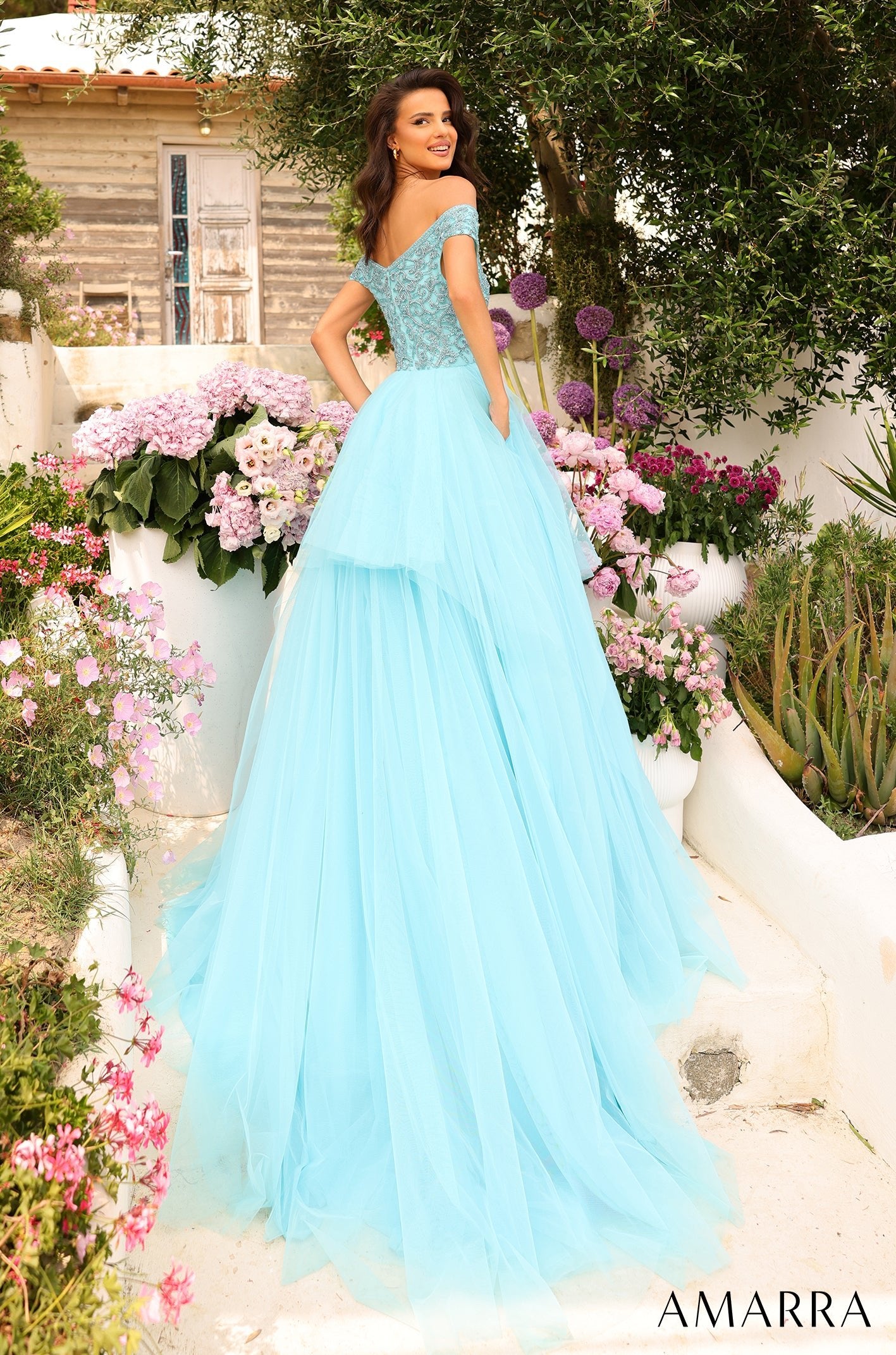 A woman in a flowing teal ball gown poses among pink and purple flowers in a garden setting.