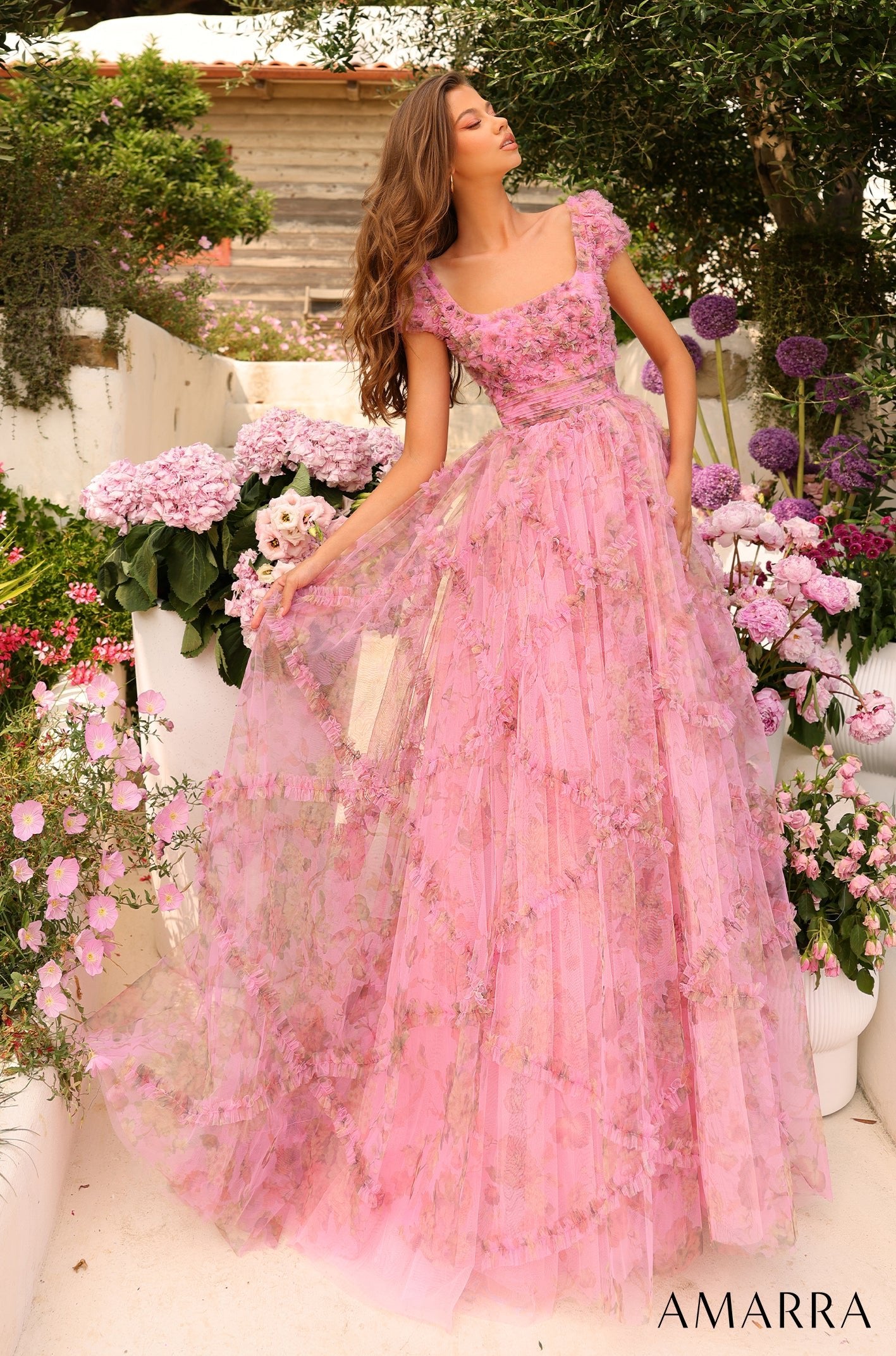A woman in a voluminous pink floral gown stands gracefully among pink and purple flowers in a garden.