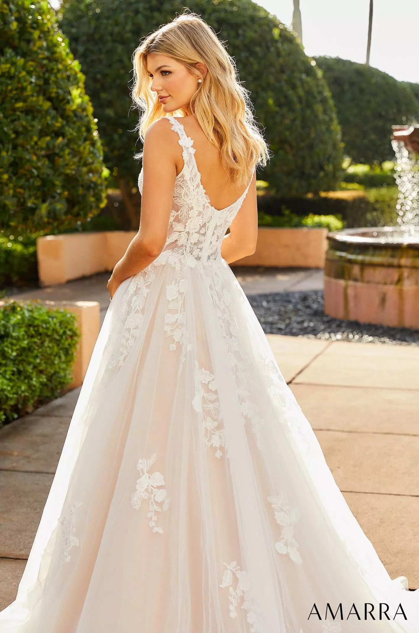A blonde woman wears a white lace wedding gown with floral appliqués in a garden setting.