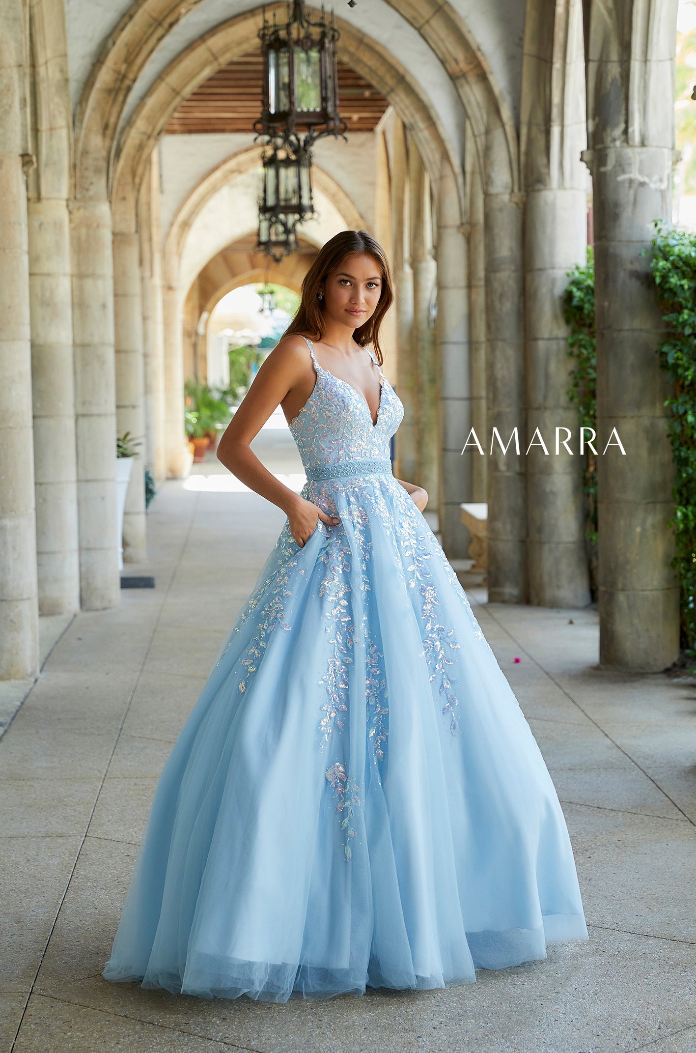 A woman wears a light blue ballgown with floral embroidery, posing under stone arches.