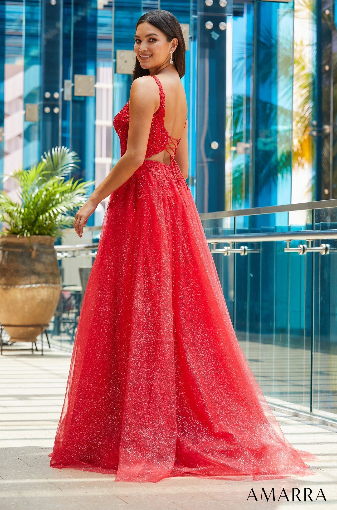 A woman wears a sparkly red floor-length gown with a corset-style back in a modern glass-walled setting.