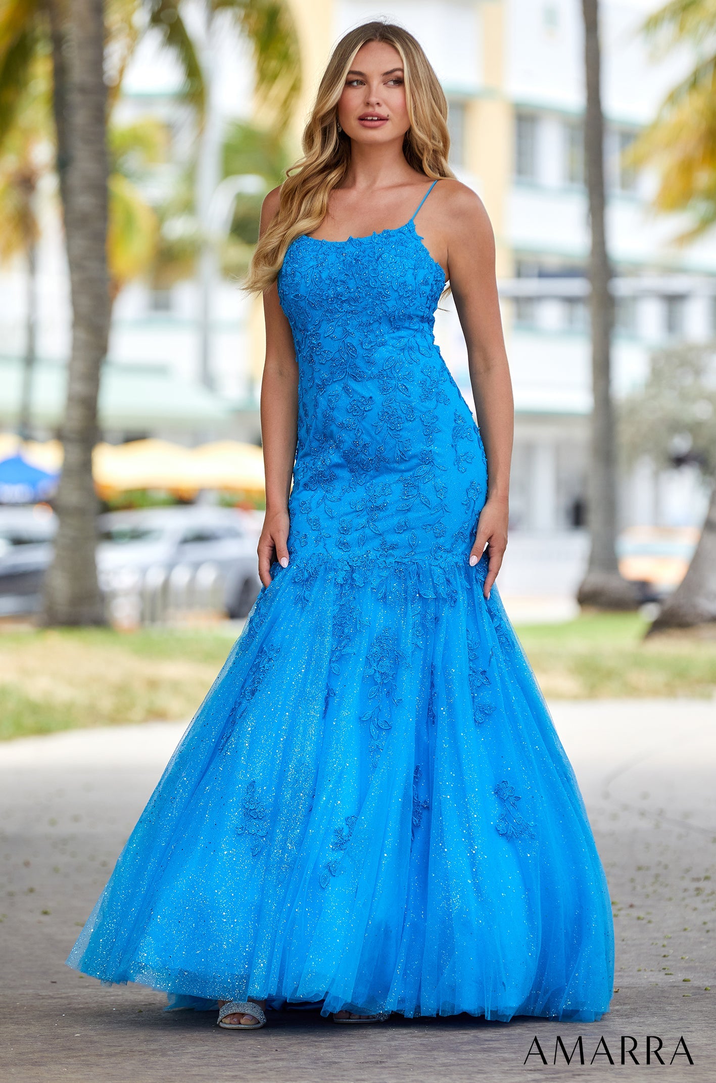 A woman wears a vibrant blue lace mermaid gown with a sparkly tulle skirt against a palm tree backdrop.