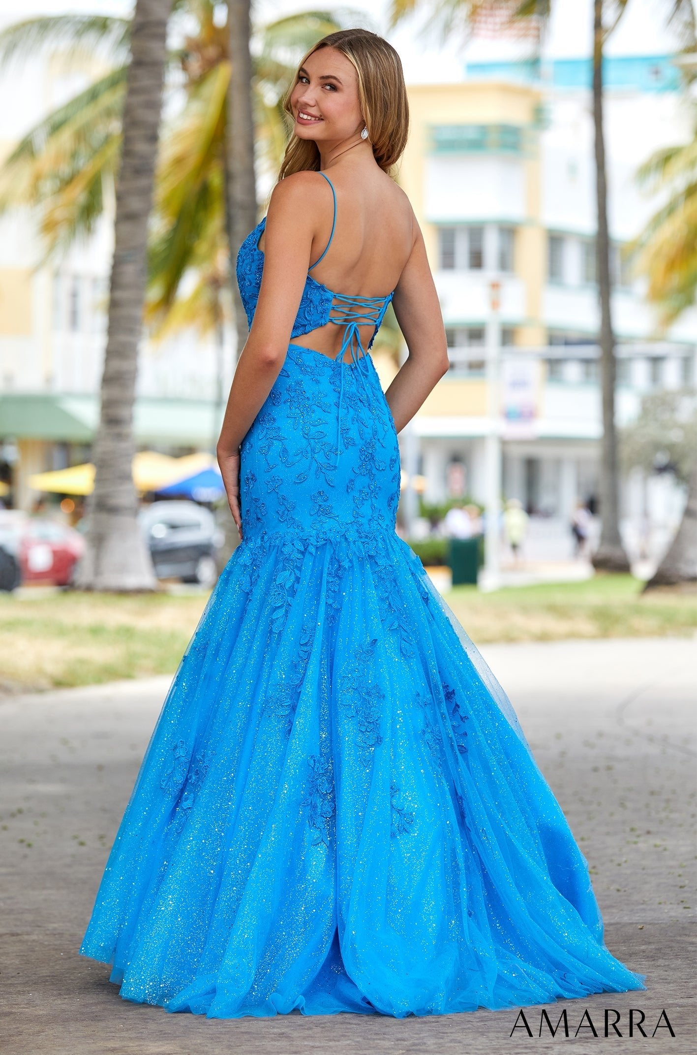 A vibrant blue mermaid-style gown with a corset back, worn against a palm tree-lined urban backdrop.