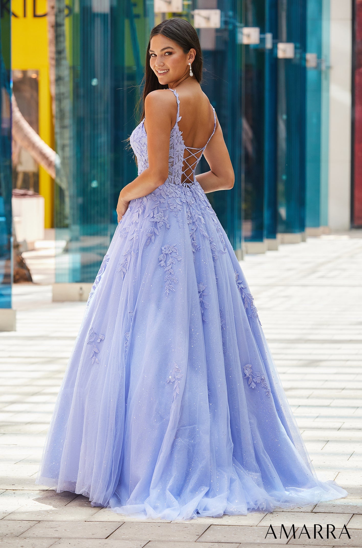 Lavender ball gown with lace appliques and a corset back, posed on a urban walkway with glass walls.