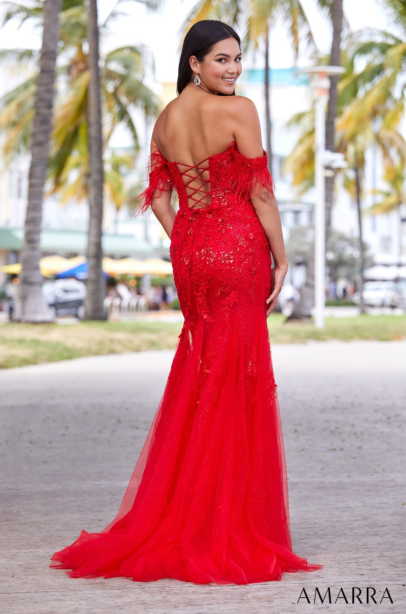 A woman wears a vibrant red sequined gown with feathered trim and a corset back, posing among palm trees.