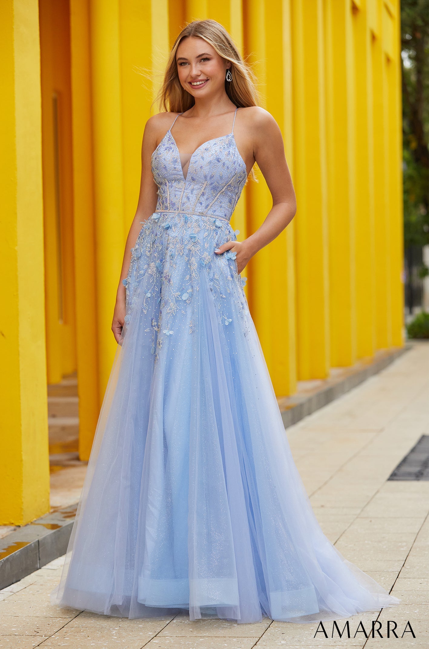 A light blue tulle A-line gown with floral embellishments and spaghetti straps stands against a yellow background.
