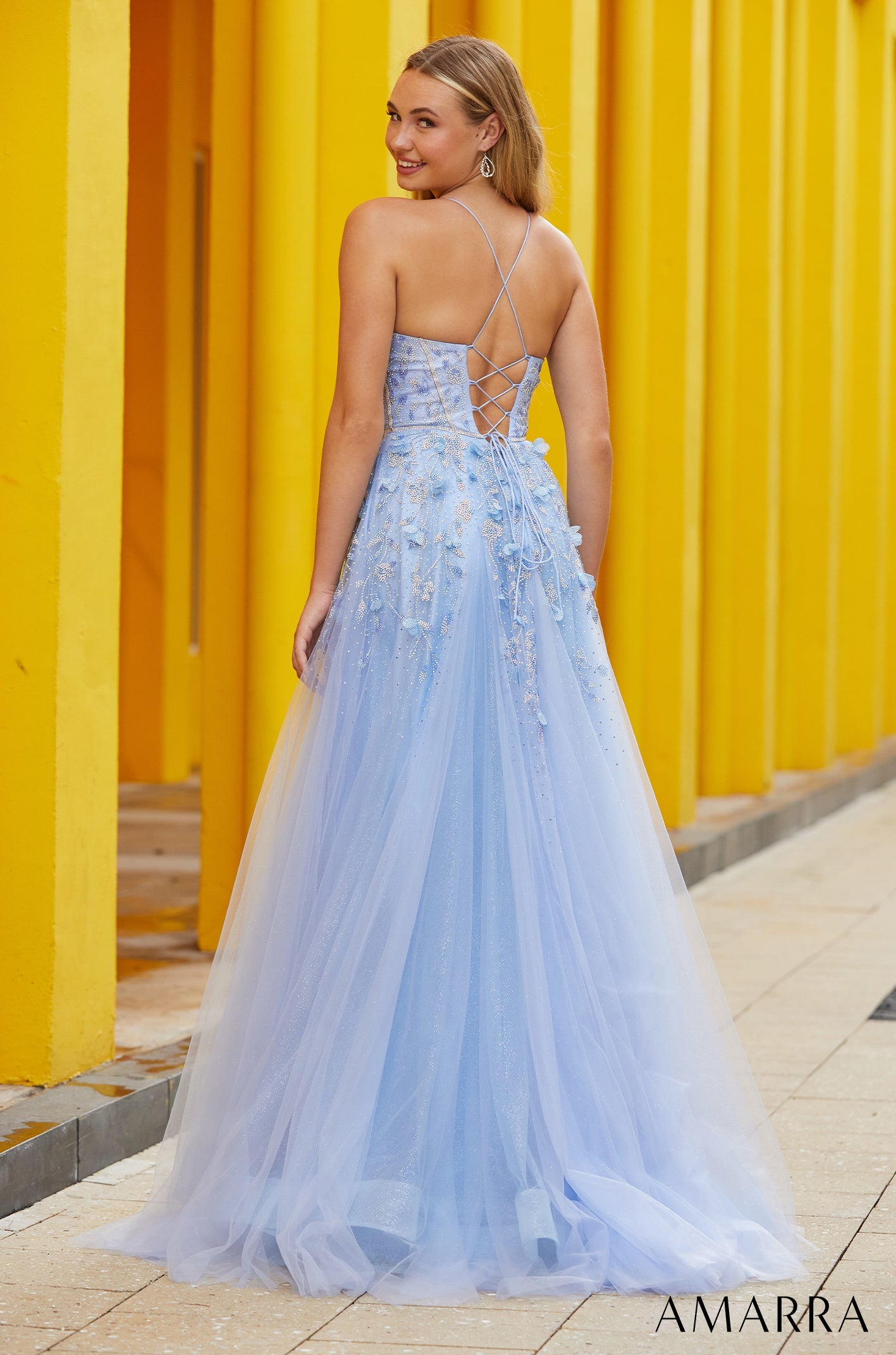 A light blue tulle gown with floral embellishments and a criss-cross back, photographed against a yellow striped wall.