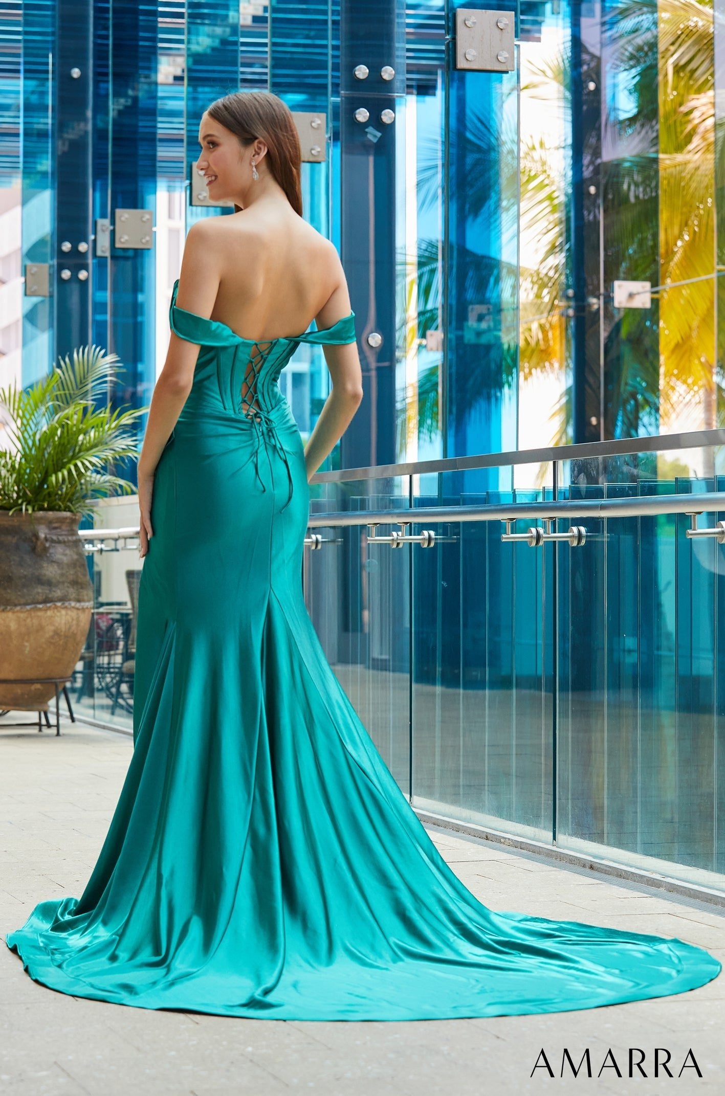 A teal mermaid-style off-shoulder gown with a dramatic train poses against a blue glass backdrop.