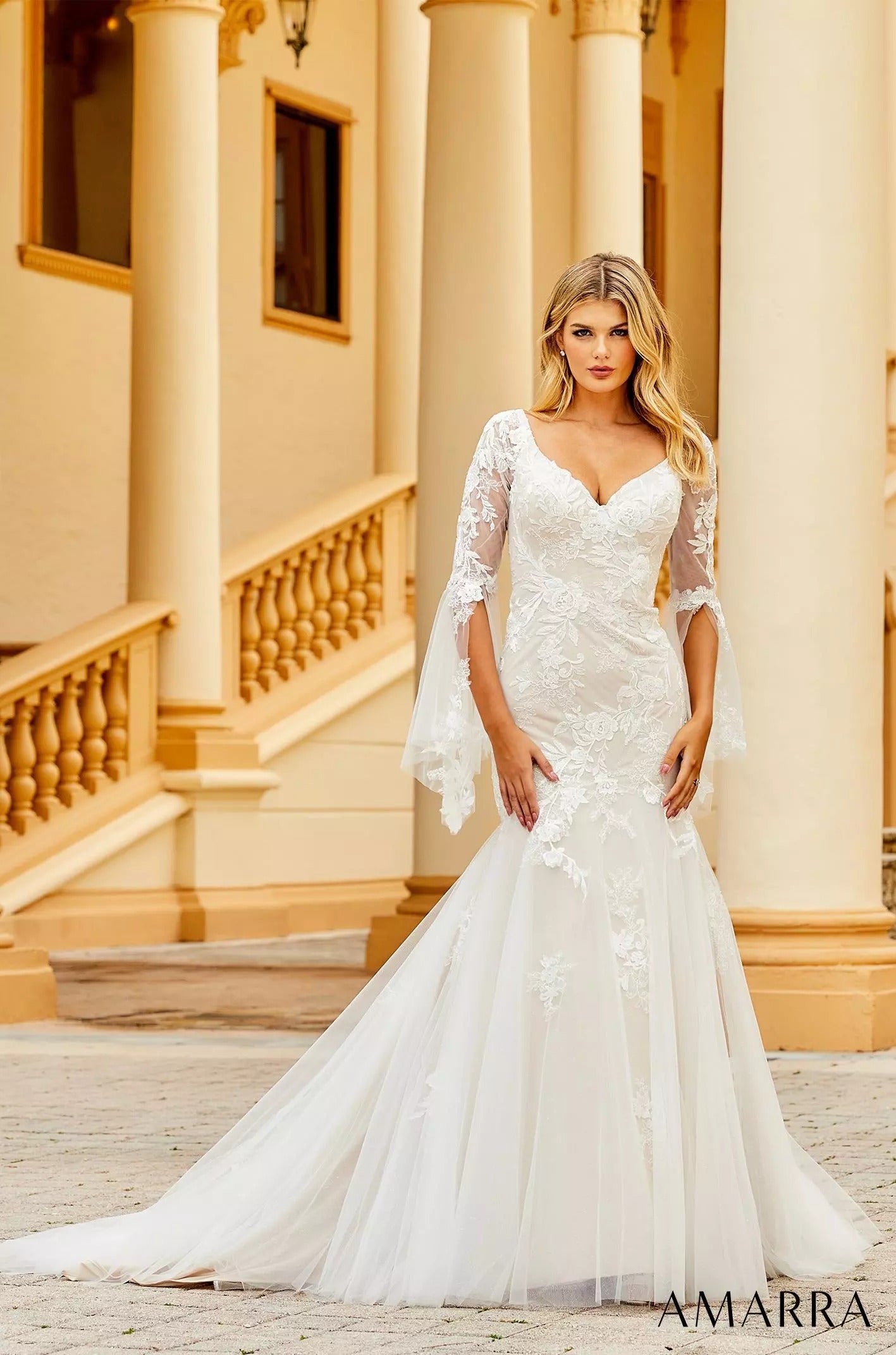 A blonde woman wears a white lace mermaid wedding dress with long sleeves in front of classical columns.