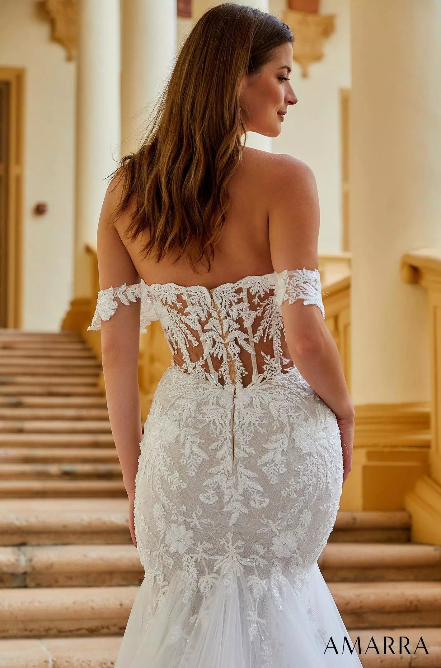 A bride in an off-shoulder lace mermaid wedding dress stands on stairs, viewed from behind.