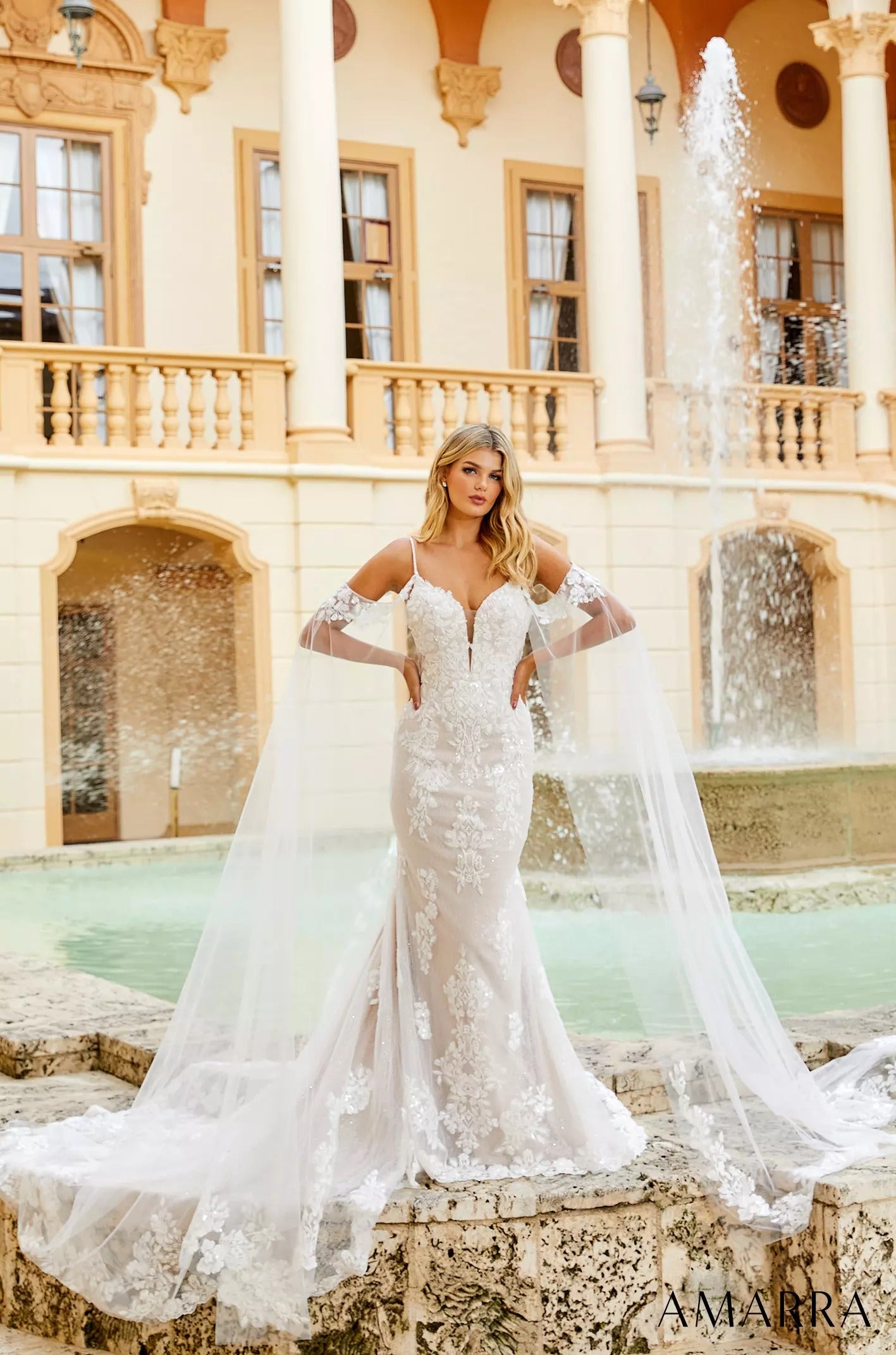 A bride in a white lace mermaid wedding dress stands by a fountain in an elegant courtyard with ornate architecture.