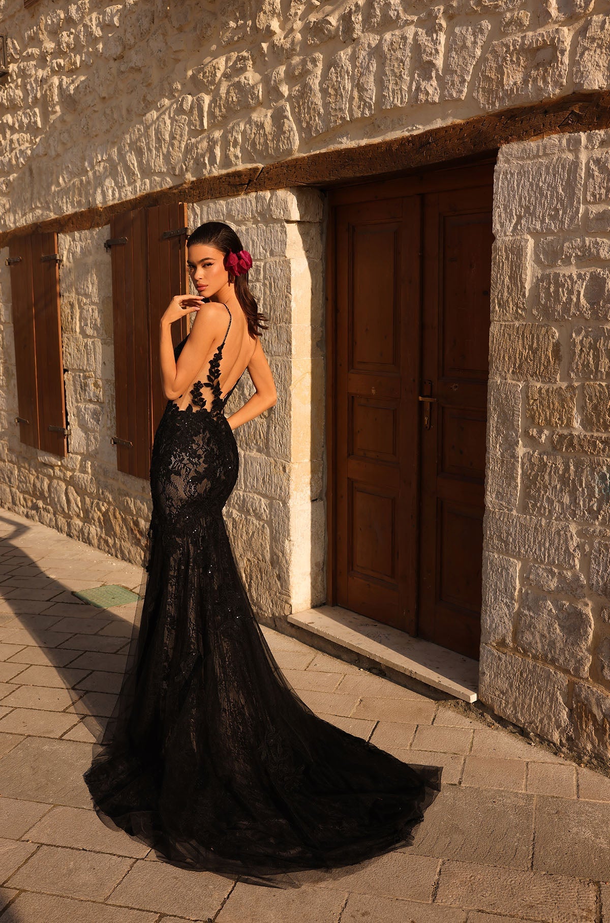 A woman in a black lace mermaid gown with a long train stands against a stone wall at sunset.