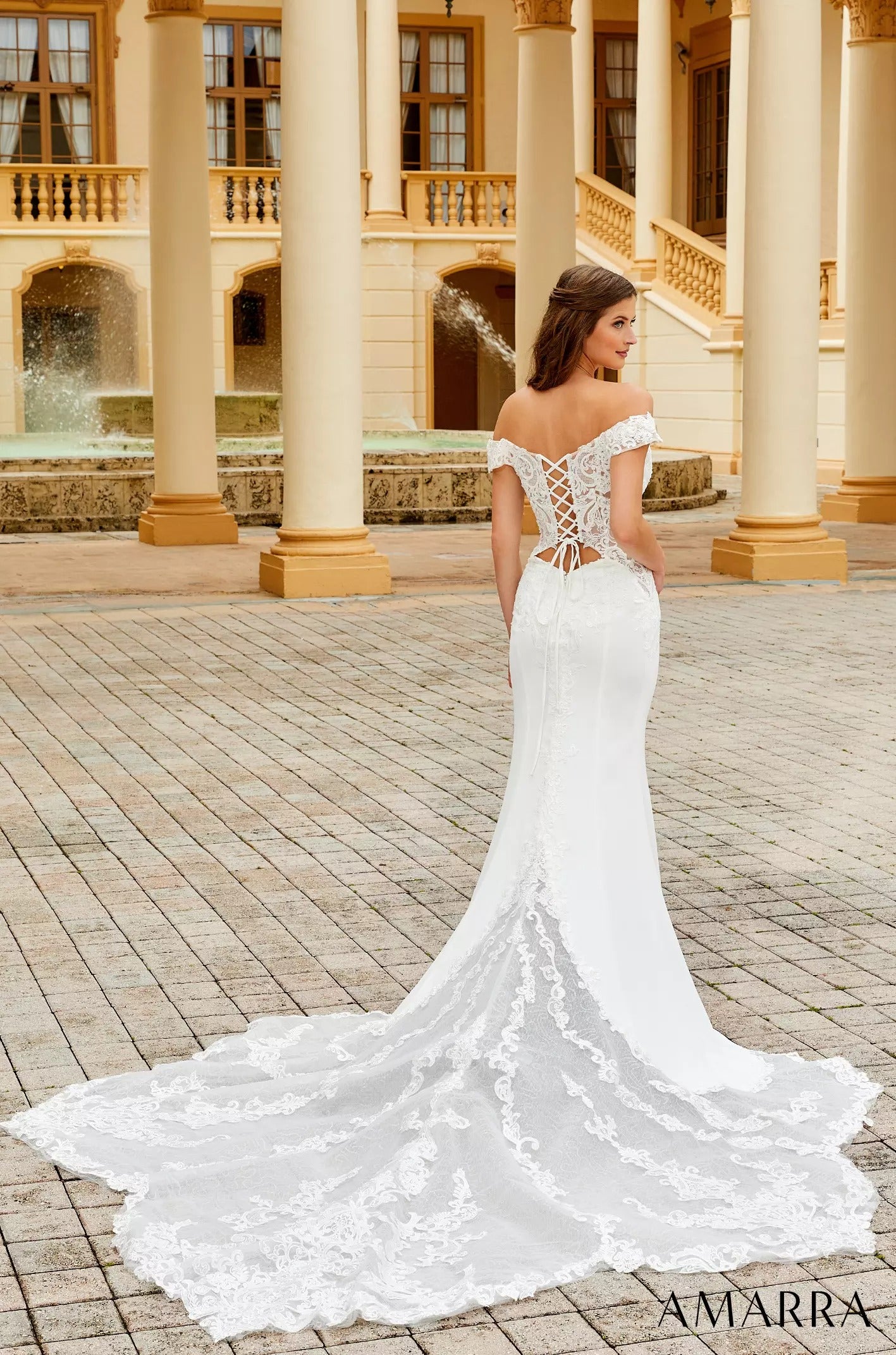 A white lace wedding dress with corset back and long train, worn by a model posing in a columned courtyard.
