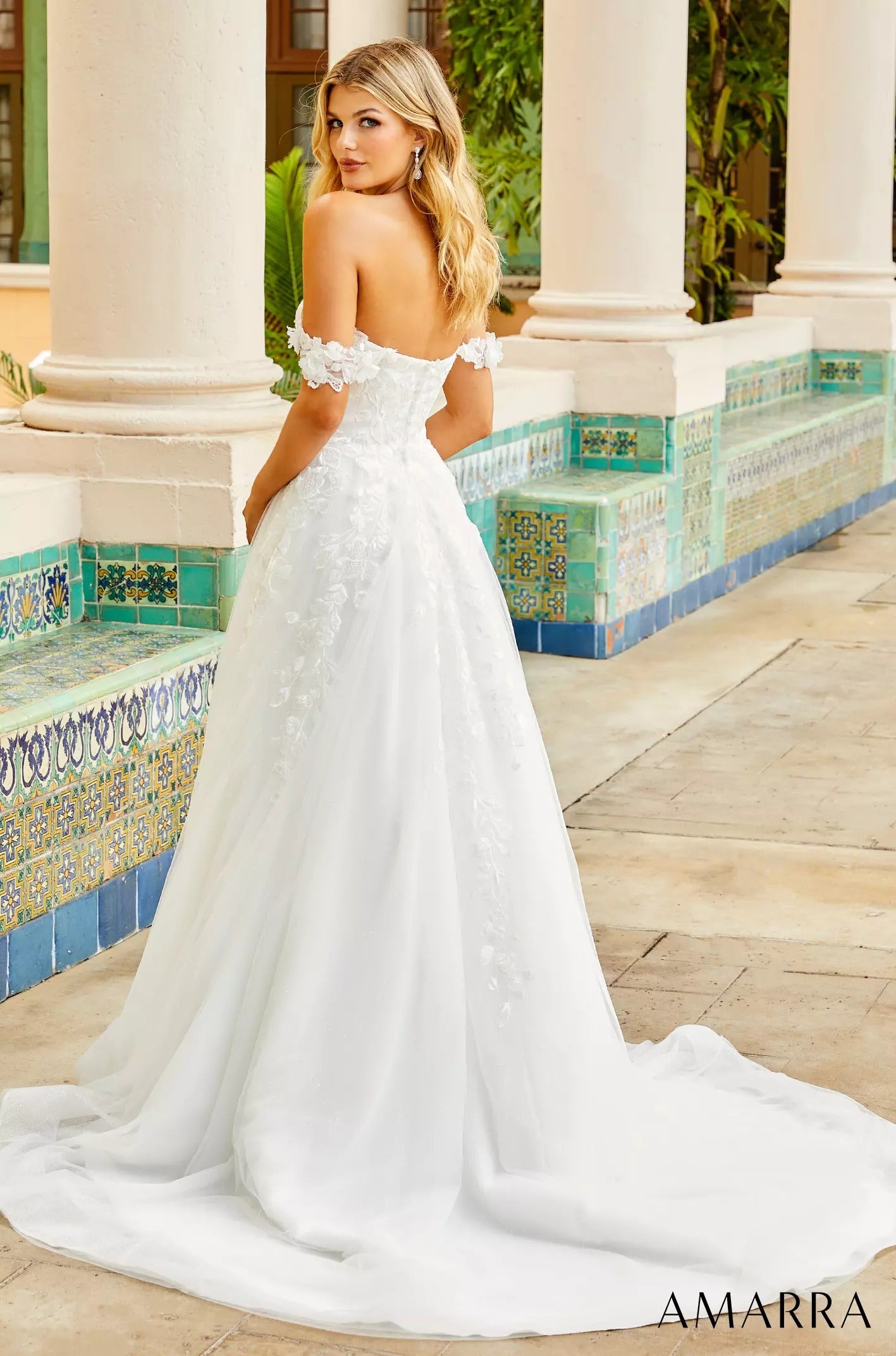 A blonde woman in an off-shoulder white wedding gown poses beside ornate blue-green tiled steps.