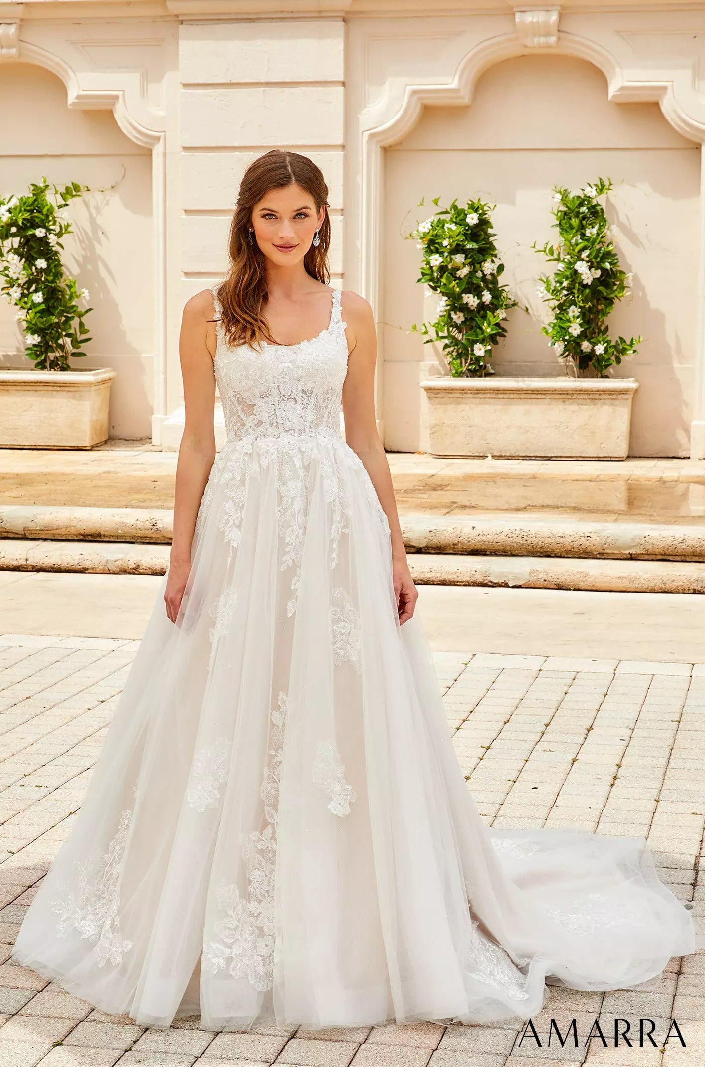 A white lace wedding gown with a full ballgown skirt, worn by a model on stone steps near potted plants.