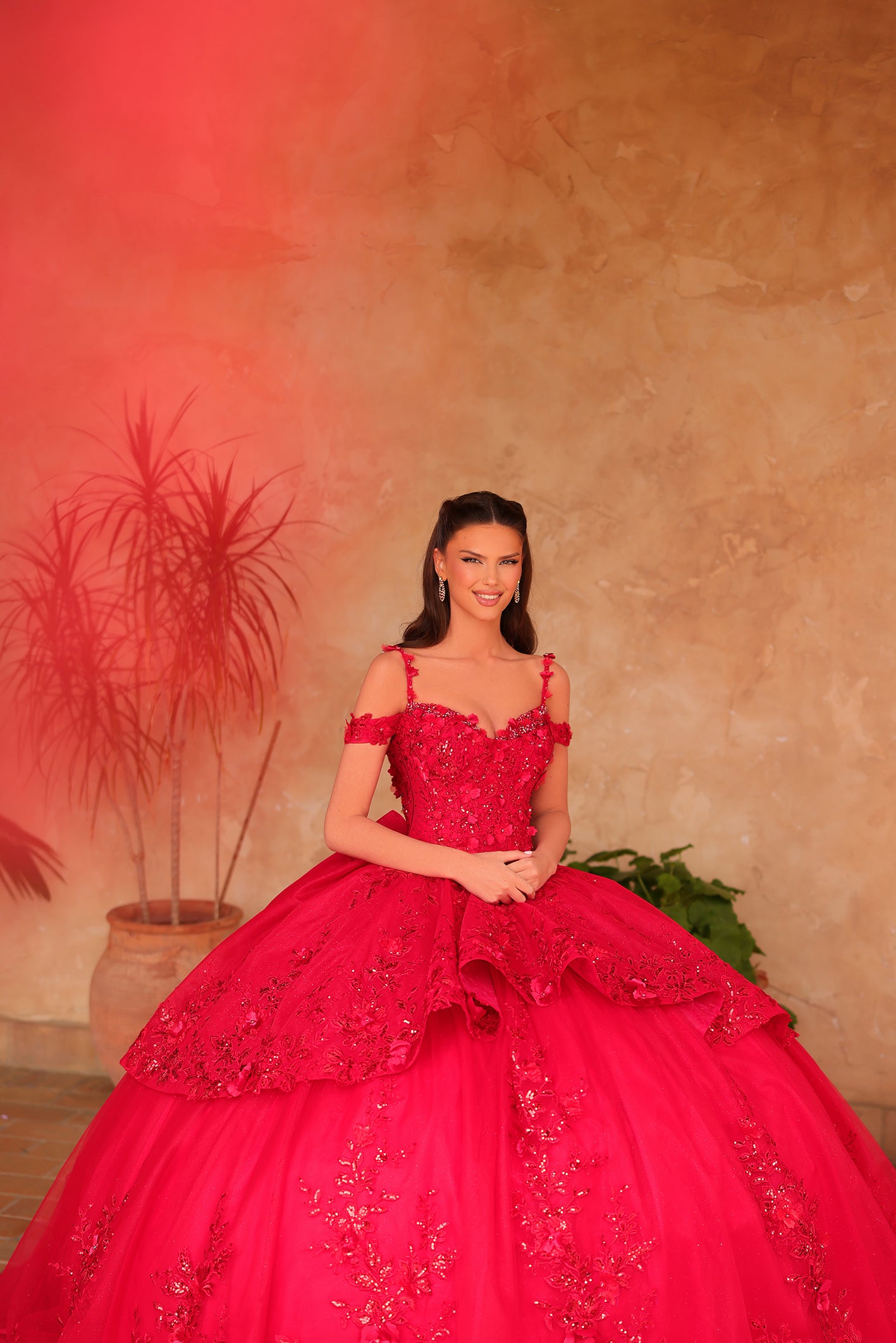 A woman in a vibrant red quinceañera ball gown poses with a warm-toned textured background and plants.
