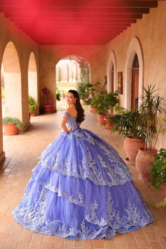 A woman in a lavish periwinkle quinceañera gown poses in an arched terracotta courtyard with plants.