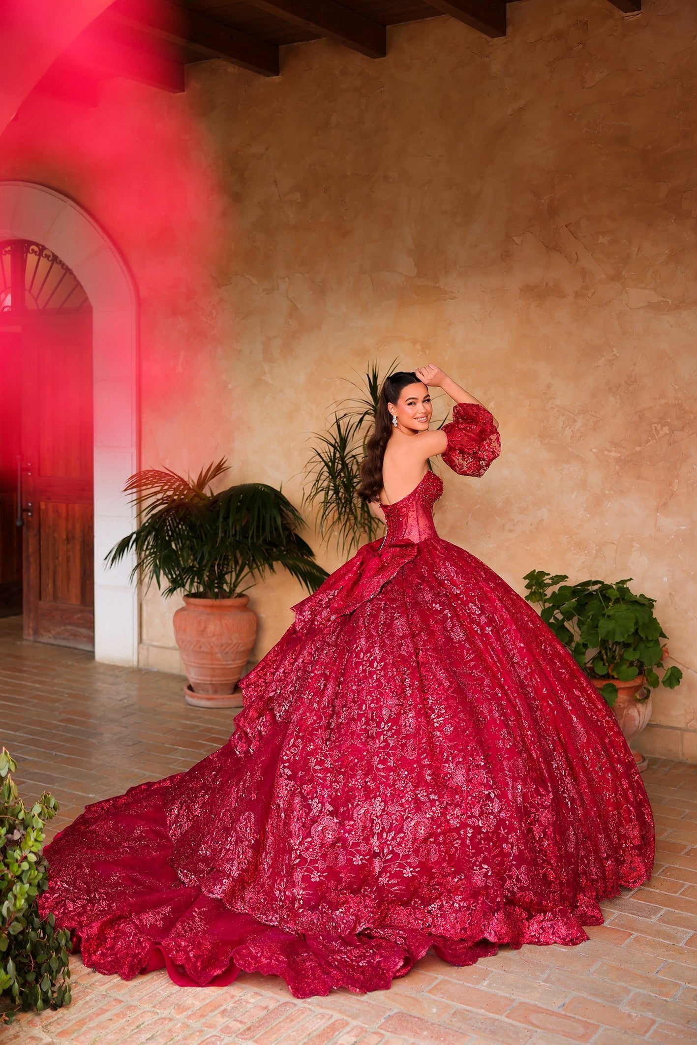 A woman in a vibrant red ball gown poses dramatically in a rustic terracotta-walled courtyard with potted plants.