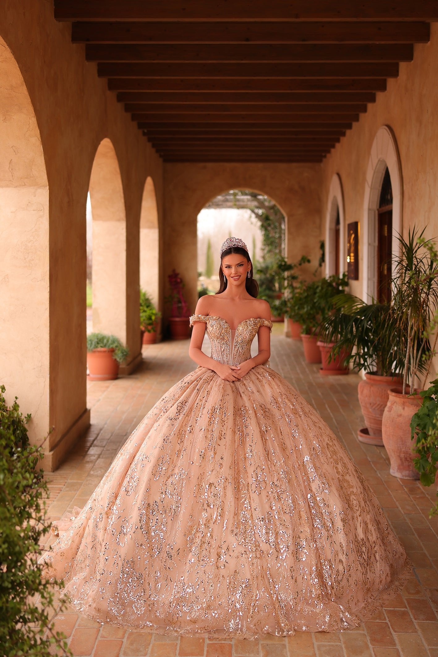 A woman wears a sparkling gold ball gown with a tiara, standing in an arched Mediterranean-style hallway.