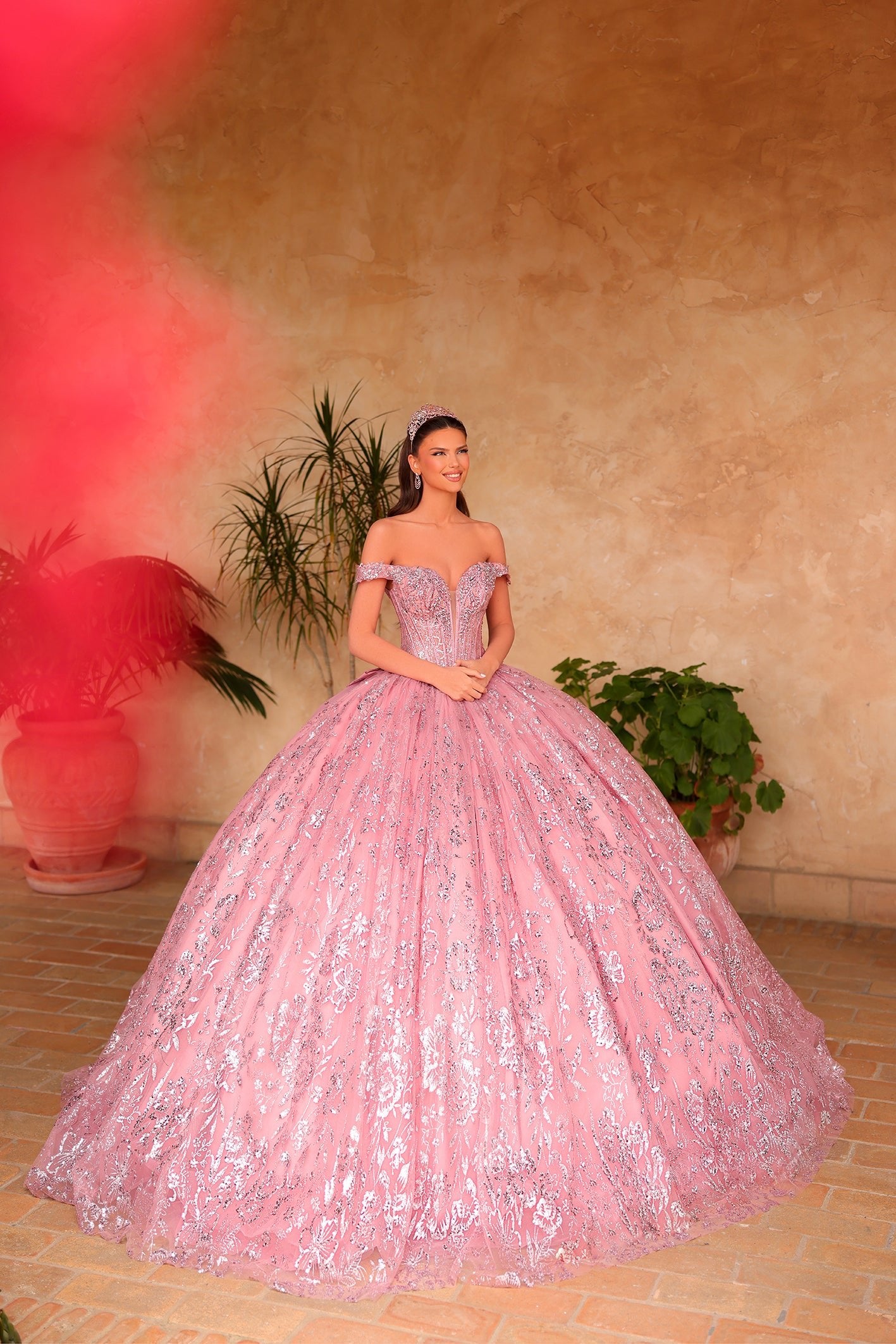A woman in a rose gold sparkly ball gown poses in a sunlit room with potted plants.