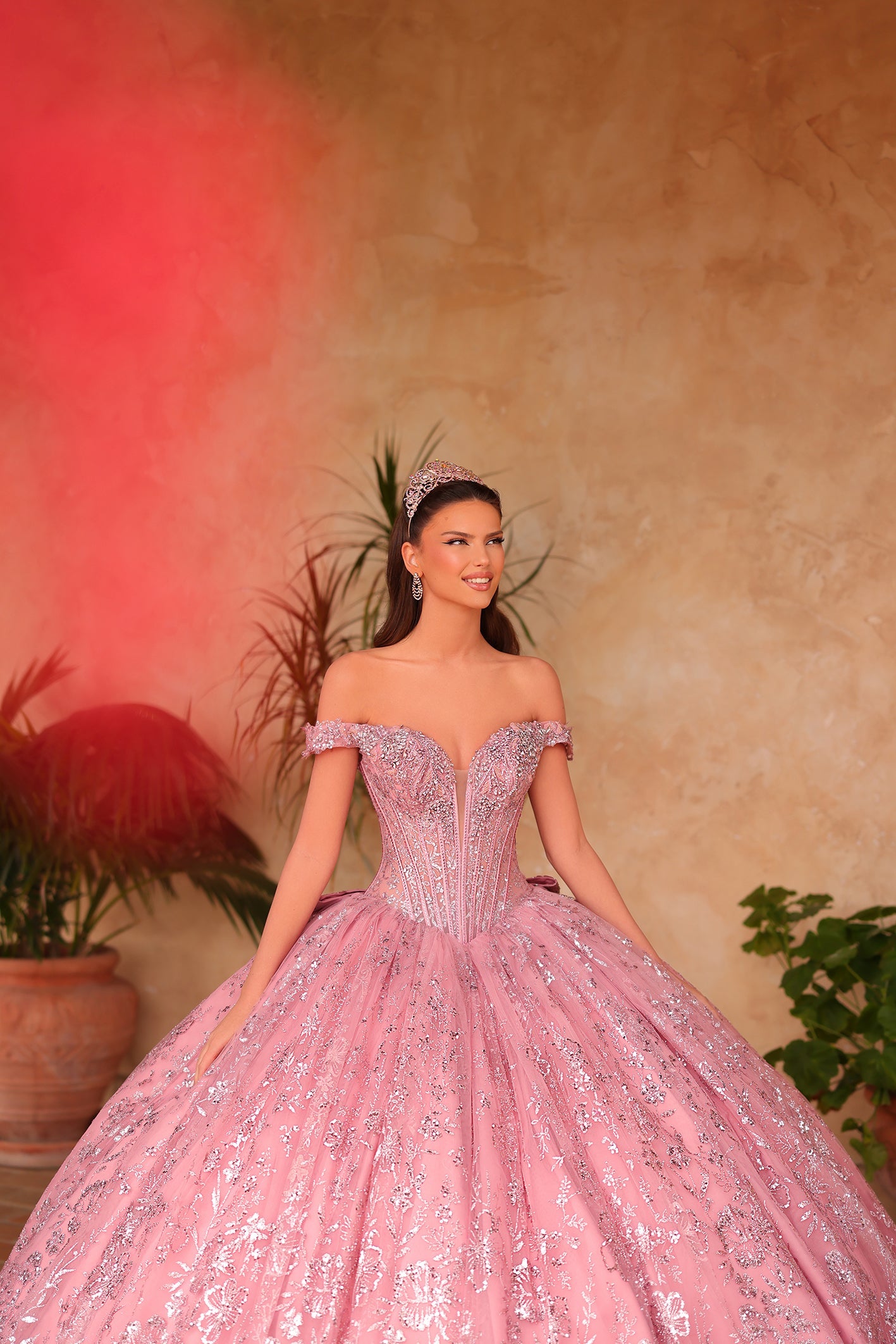 A woman wears a sparkling pink ball gown with a corset top and tiara against a warm-toned background.