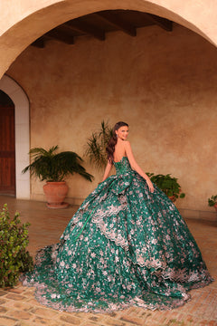 A woman in an emerald green floral ballgown poses dramatically in an arched, terracotta-walled courtyard.