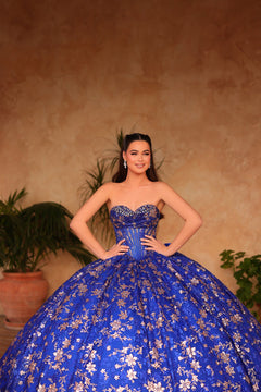 A woman wears an elaborate royal blue ball gown with floral embroidery, posing confidently against a warm background.