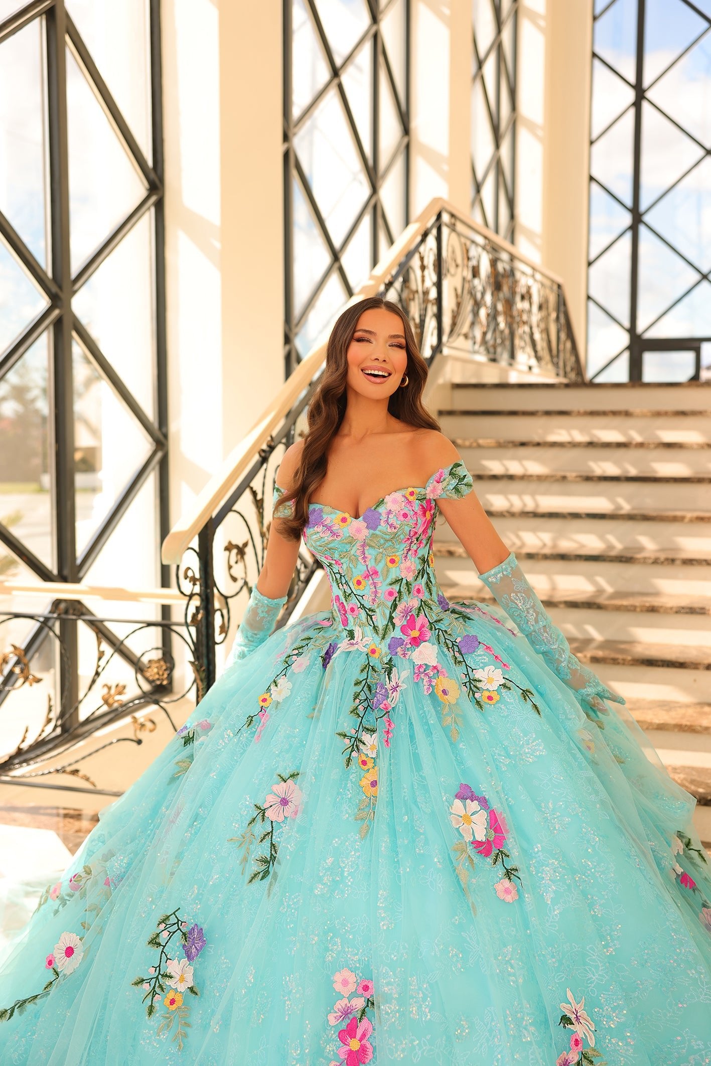 A woman in a turquoise floral ball gown smiles on an ornate staircase with sunlight streaming through windows.