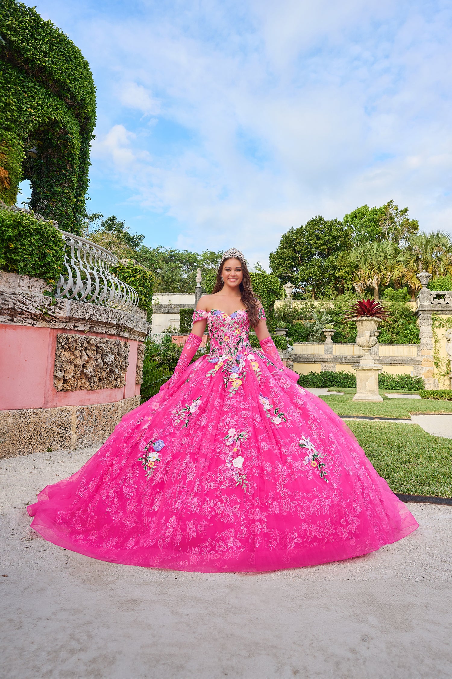 A young woman wears a voluminous Fuchsia/Multi floral quinceañera gown in an elegant garden setting.