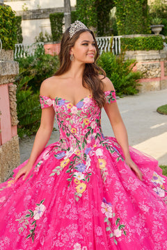 A young woman in a vibrant pink floral quinceañera gown wears a tiara and stands in a garden.