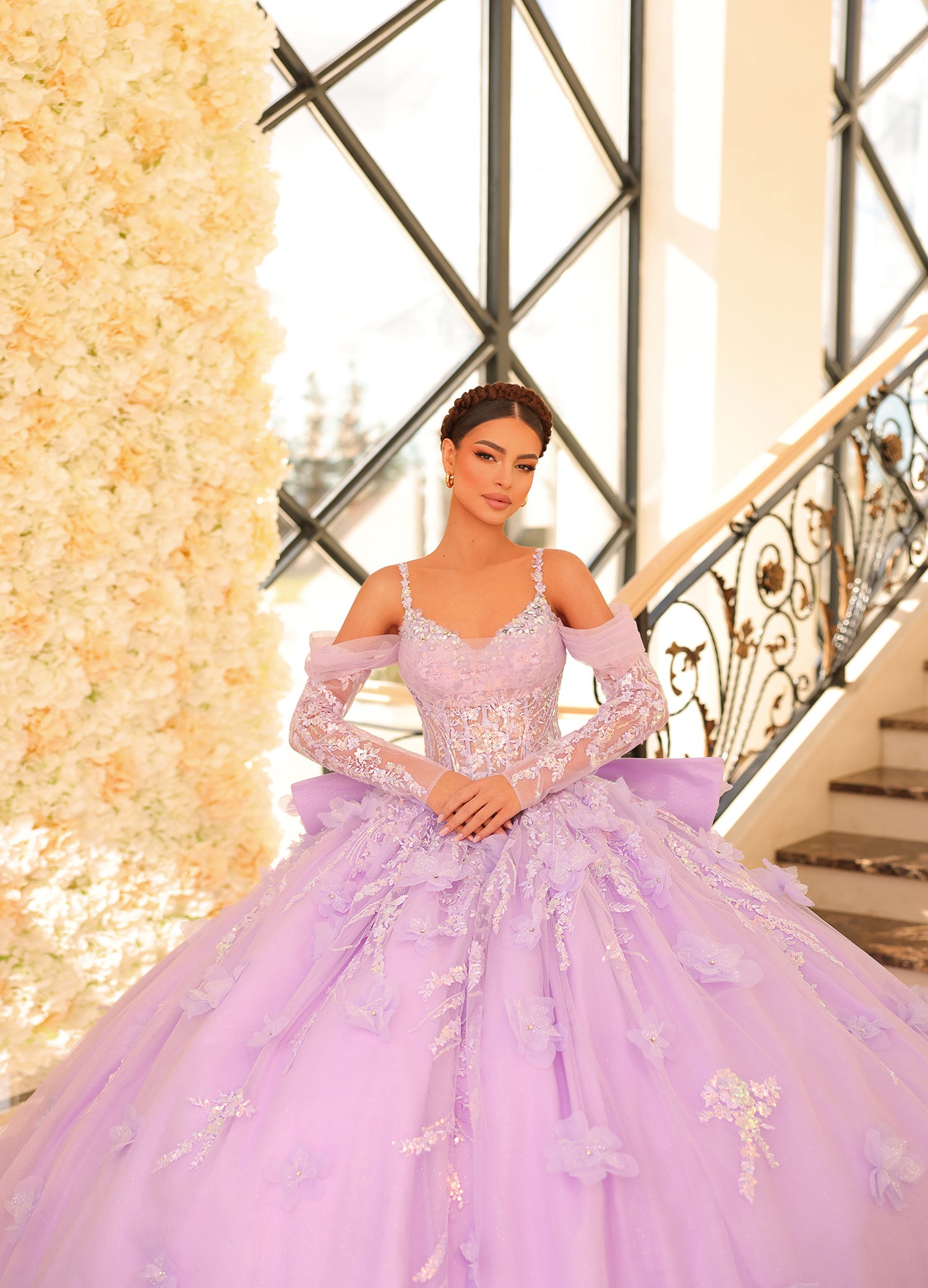 A woman in a lavender ballgown with lace sleeves poses elegantly on a staircase with floral decor.