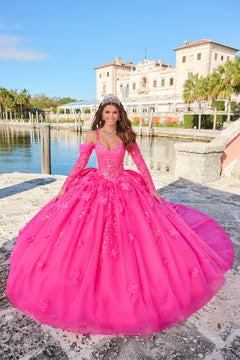 A woman wears a voluminous Hot Pink ballgown with floral accents, standing by a waterfront with a grand building behind.