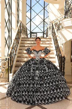 A woman stands on marble stairs wearing an elaborate Black/White floral quinceañera ball gown with off-shoulder sleeves.