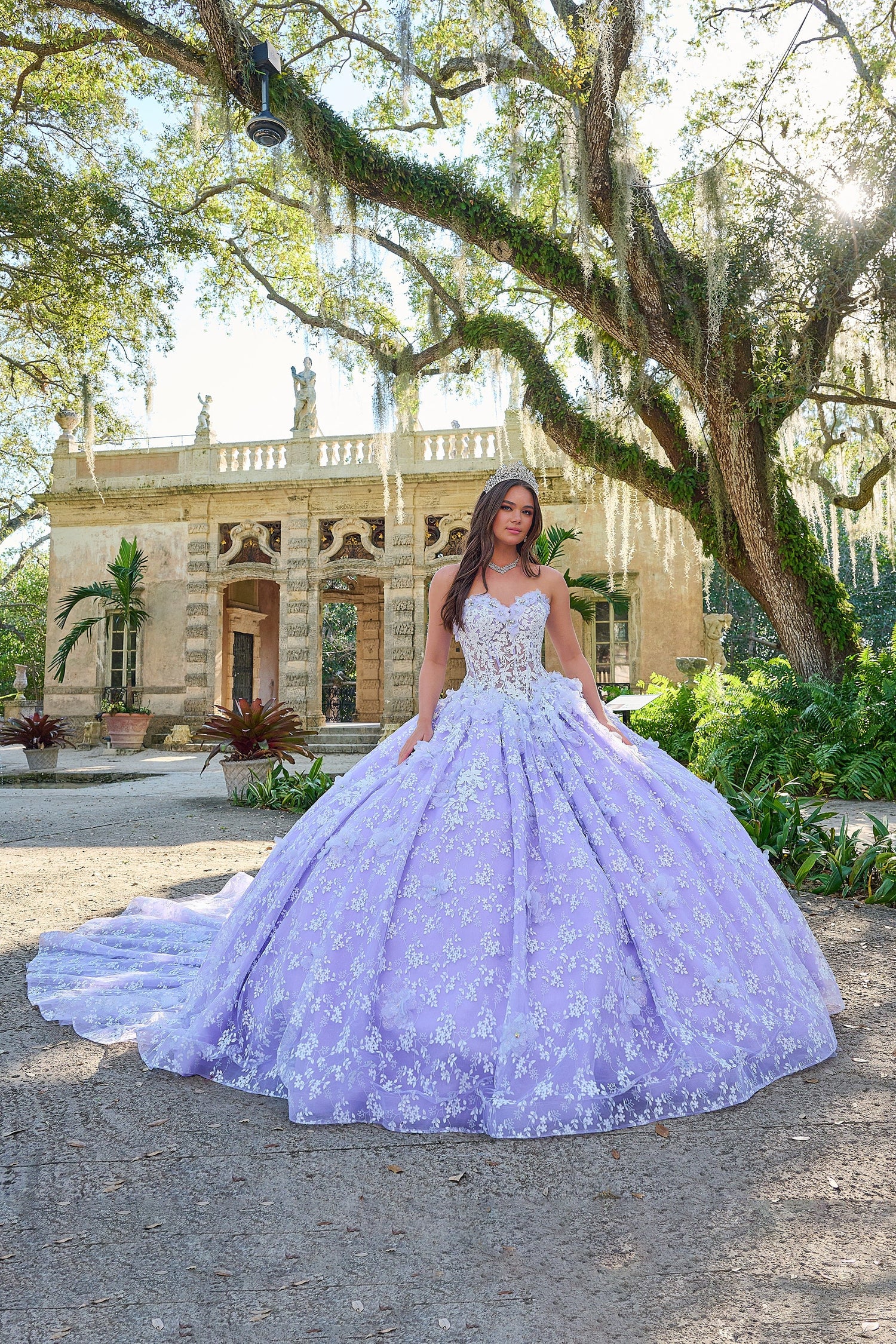 A quinceañera in a lavender ballgown poses before a historic stone building with moss-draped trees.