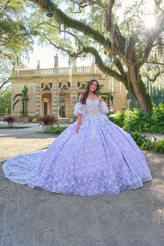 A young woman in a lilac quinceañera gown poses in a lush garden with a historic stone building behind her.