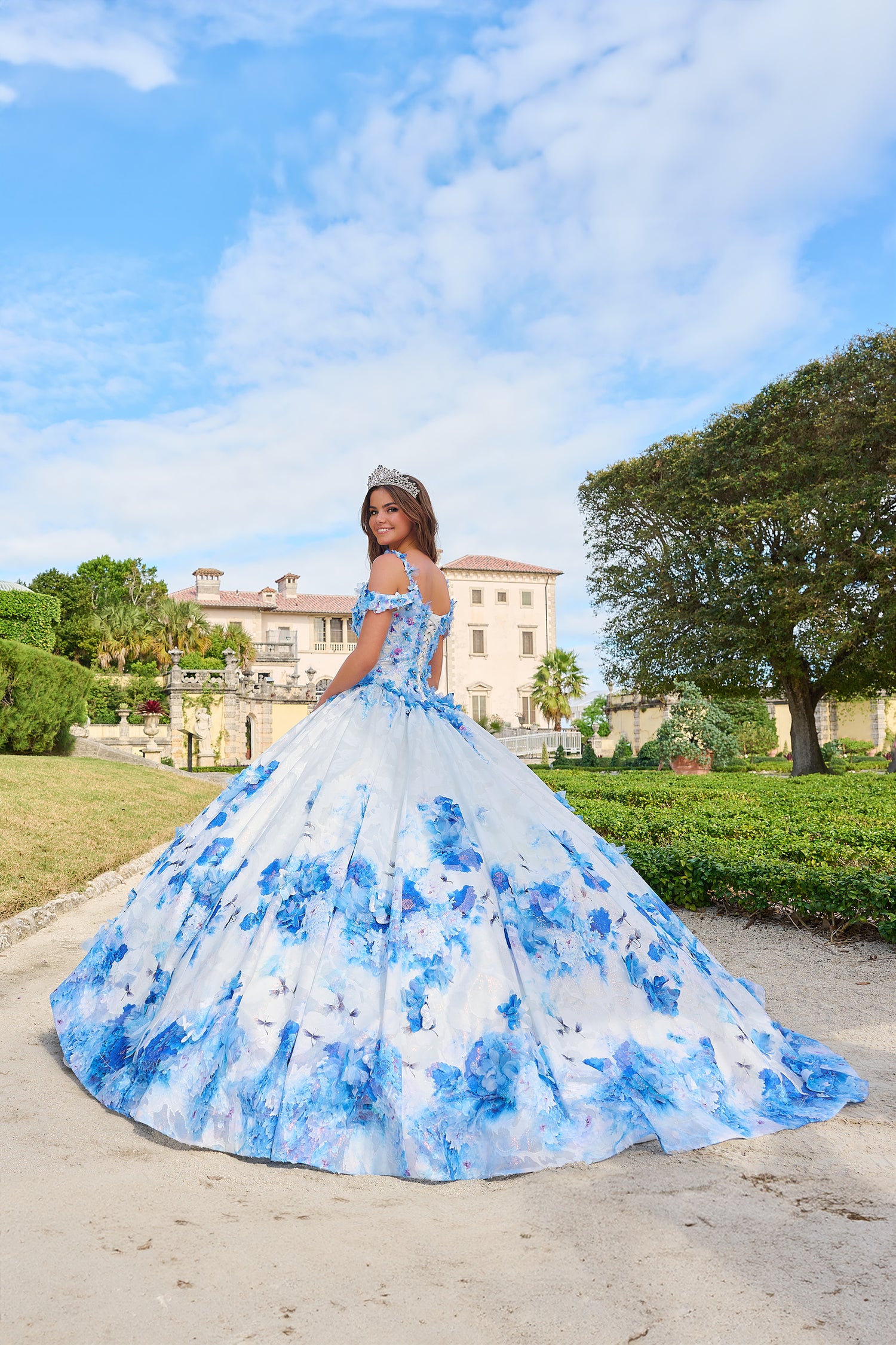 A woman in a blue and white floral quinceañera gown stands in a garden with a historic mansion behind her.