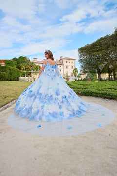 A woman in a blue floral ball gown poses in a garden with a large mansion in the background.