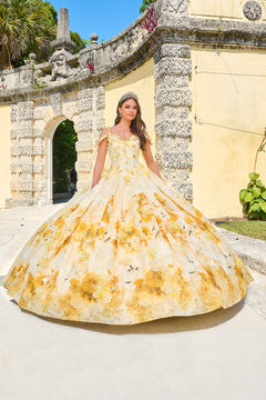 A young woman wears a voluminous Yellow floral ball gown near a stone archway with lush greenery.