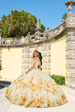 A young woman in a yellow floral ball gown poses at an ornate stone wall with palm trees and blue sky.