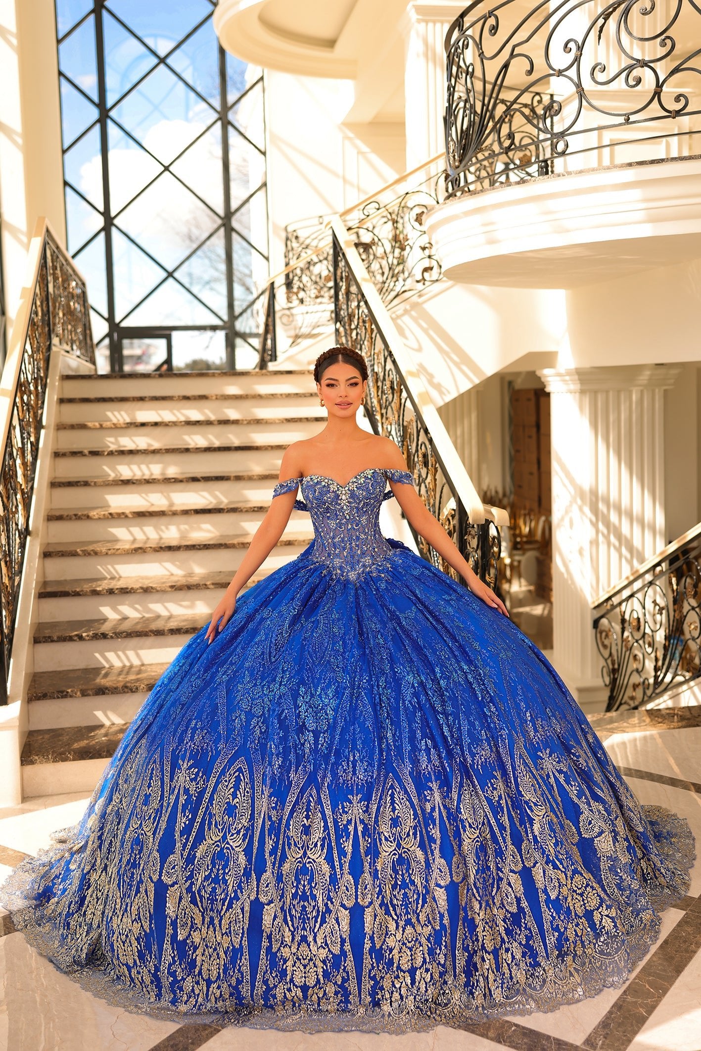 A woman in a royal blue off-shoulder ball gown stands on an ornate staircase with intricate metalwork.