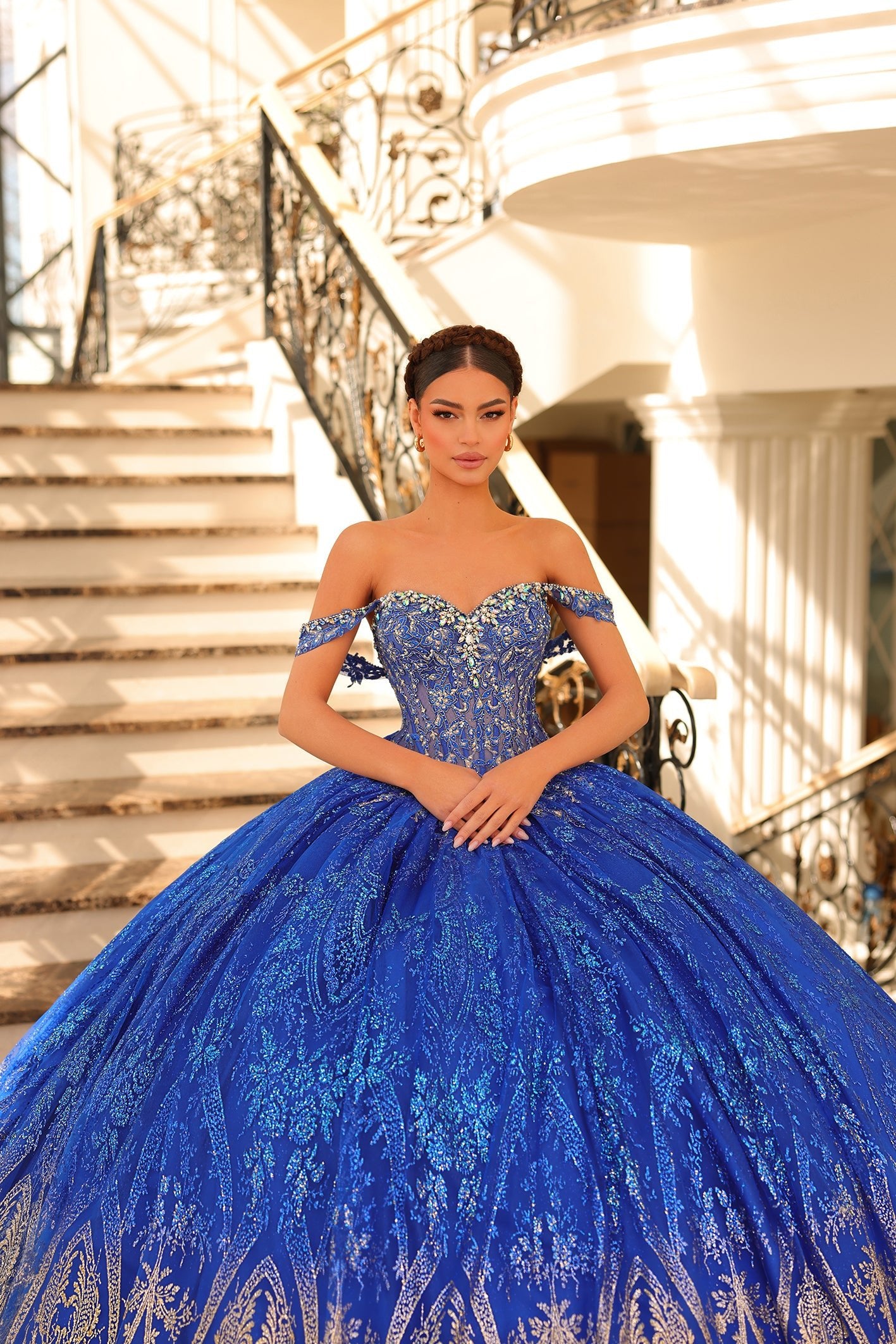 A woman in a royal blue, off-shoulder ball gown stands on an elegant staircase with ornate railings.