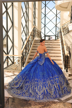 A woman in a royal blue ballgown with gold embellishments poses on an elegant staircase with ornate railings.