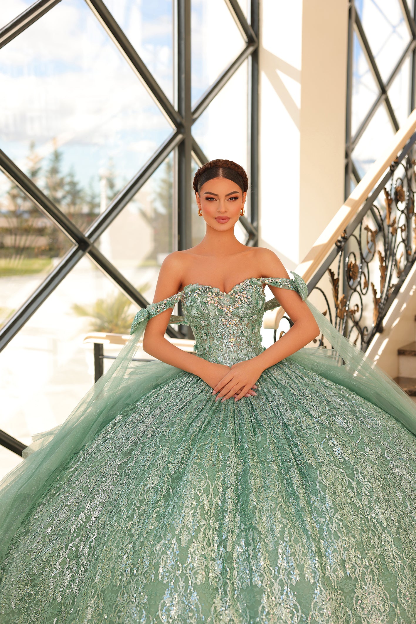 A woman in a sage green sequined ballgown stands by a window with ornate metalwork.