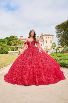 A young woman in a vibrant red ball gown with a tiara stands before an elegant estate and manicured gardens.