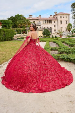 A woman in a vibrant red ball gown and tiara poses in a manicured garden with an elegant mansion backdrop.
