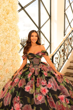 A woman wears an off-shoulder floral ball gown with a corset bodice, posing on an elegant staircase.