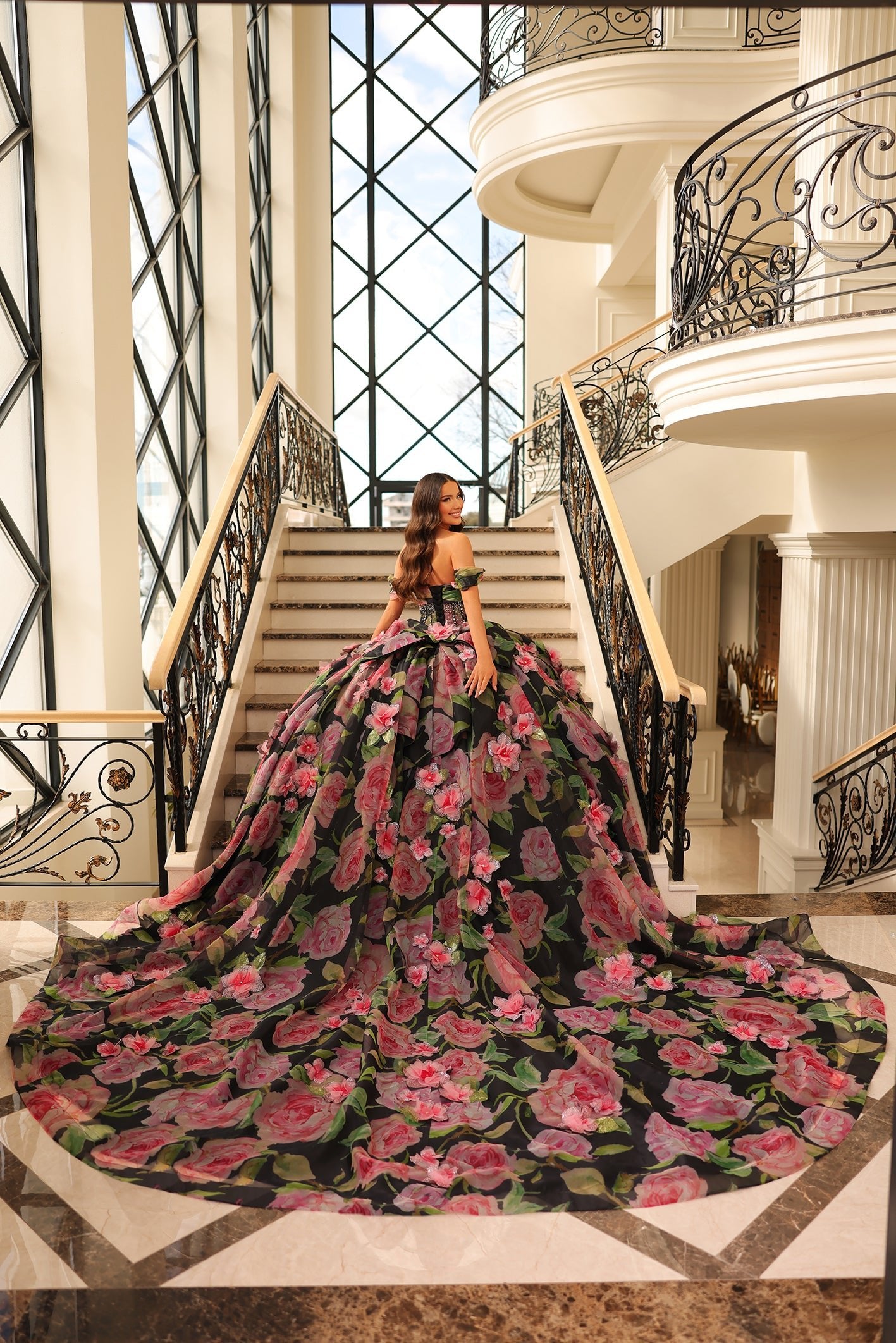 A woman in a floral ball gown stands on an elegant staircase with ornate iron railings and large windows.