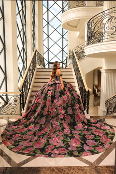 A woman in a floral ball gown stands on an elegant staircase with ornate iron railings and large windows.