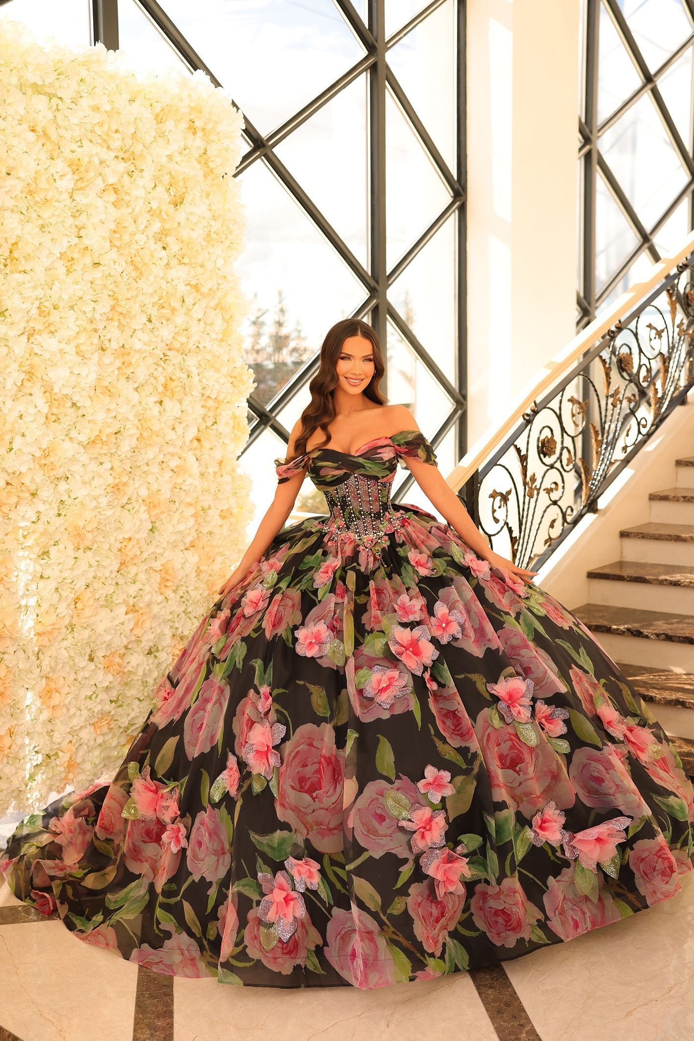 A woman in a Black/Multi floral ball gown stands by a flower wall on an elegant staircase.