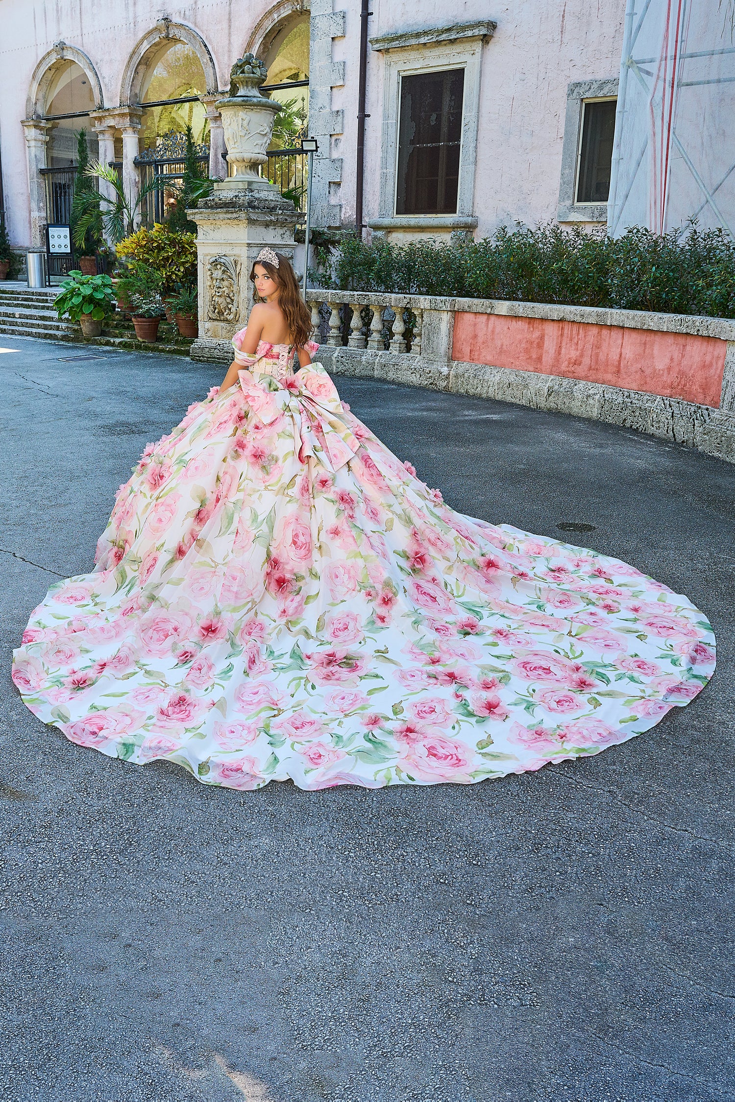 A woman wears a voluminous pink floral ballgown with a long train in front of an elegant stone building.